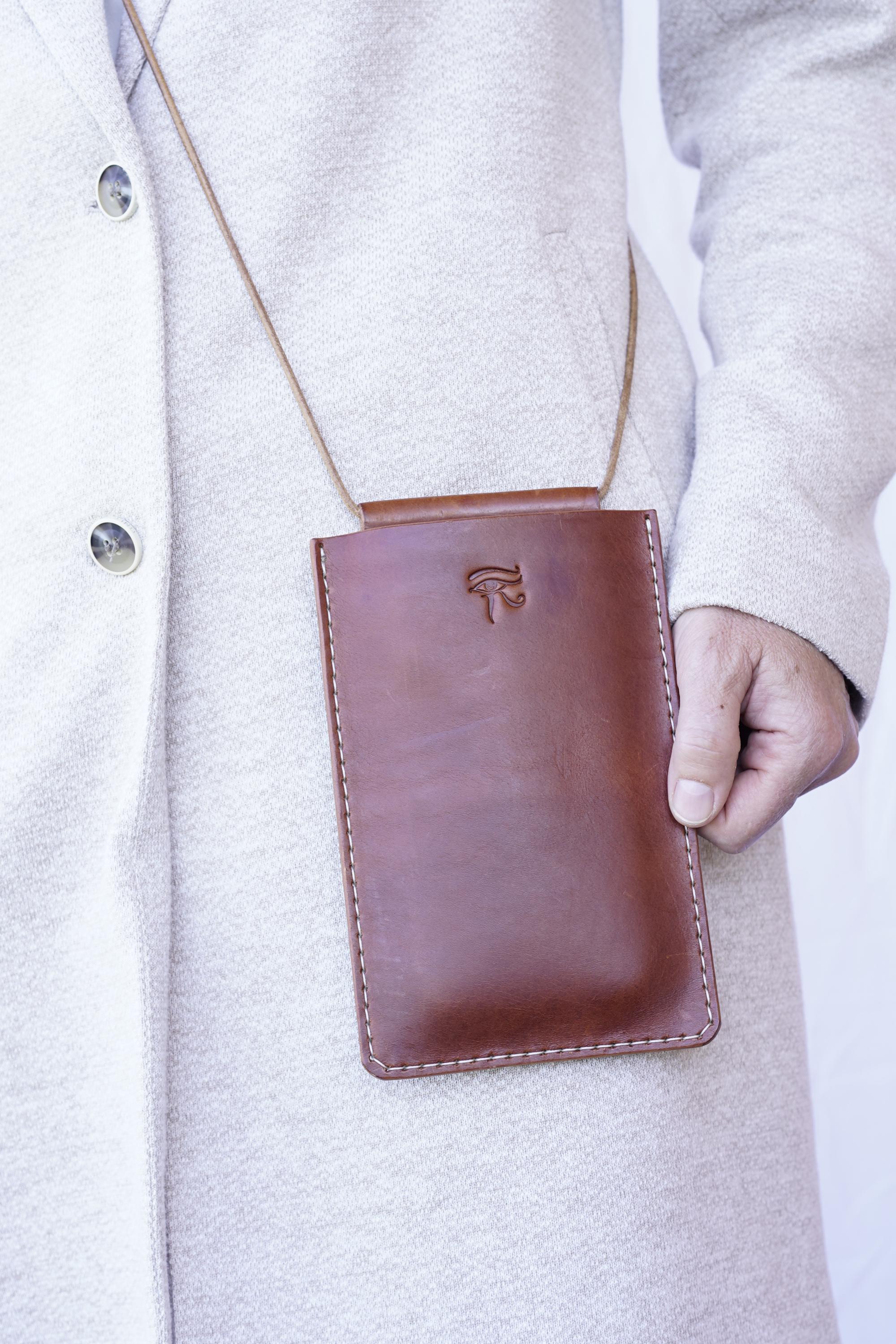 Könnte beinhalten: Eine braune Leder-Umhängetasche für ein Telefon mit einer genähten Bordüre und einem Riemen. Die Tasche hat ein geprägtes Symbol auf der Vorderseite. Die Tasche wird von einer Person getragen, die einen hellen Mantel trägt.