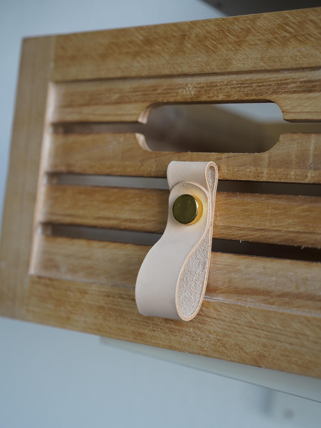 Könnte beinhalten: Nahaufnahme einer Holzschublade mit einem hellfarbenen Ledergriff. Die Schublade hat ein Lamellendesign und einen Ausschnitt für den Griff. Der Griff ist mit einem goldfarbenen Knopf befestigt.