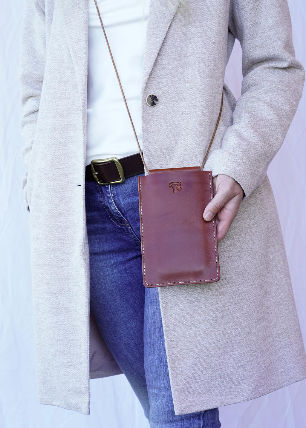 Könnte beinhalten: Eine braune Leder-Umhängetasche für das Telefon mit einer genähten Bordüre und einem kleinen geprägten Design. Die Tasche wird mit einem hellbraunen Riemen getragen. Die Person trägt einen hellgrauen Mantel, blaue Jeans und einen braunen Gürtel.