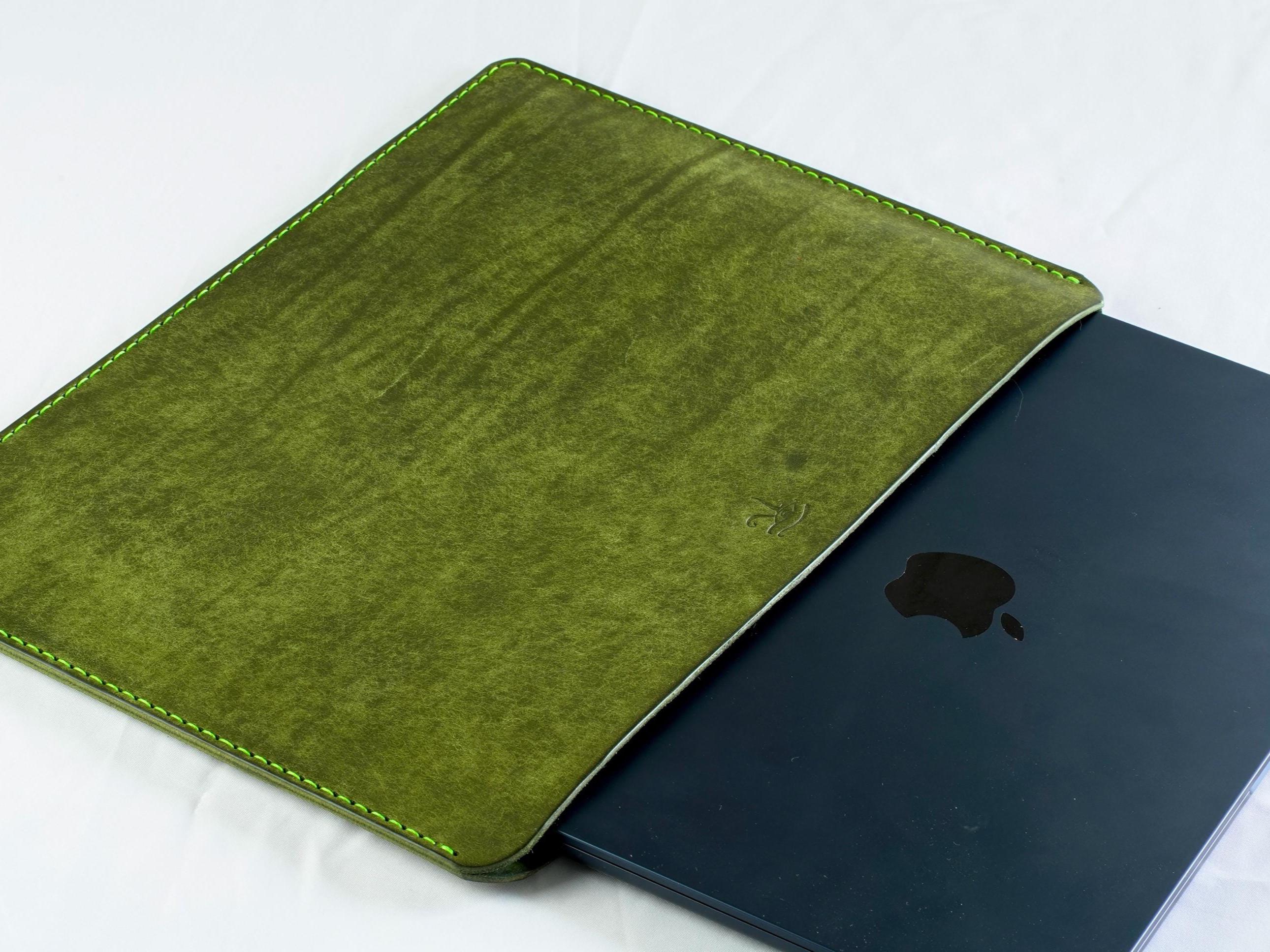 Könnte beinhalten: Eine grüne Lederhülle mit strukturierter Oberfläche, akzentuiert durch limettengrüne Nähte. Ein dunkelblauer Laptop mit dem Apple-Logo ist teilweise sichtbar. Die Hülle dient zum Schutz eines Laptops.