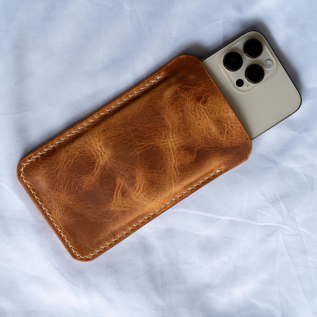 Könnte beinhalten: Eine braune Leder-Handyhülle mit weißen Nähten. Die Hülle besteht aus strukturiertem Leder und hält ein Smartphone mit einer Dreifach-Kamera. Die Handyhülle ist rechteckig und hat abgerundete Ecken. Der Hintergrund ist ein weißer Stoff.