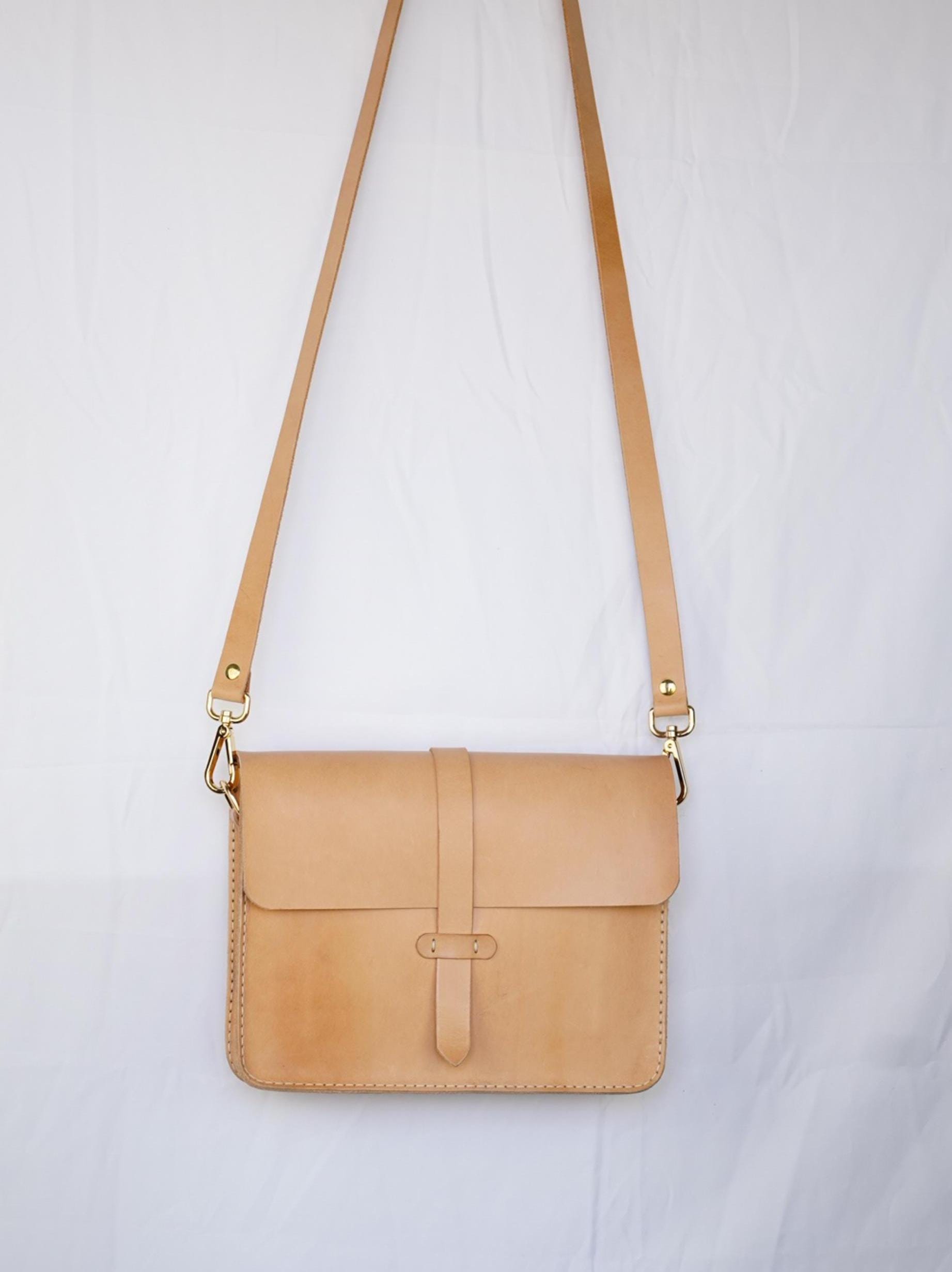 Könnte beinhalten: Eine braune Leder-Umhängetasche mit einer goldenen Schnalle und einem langen Riemen. Die Tasche hat einen Klappverschluss und ein genähtes Design.