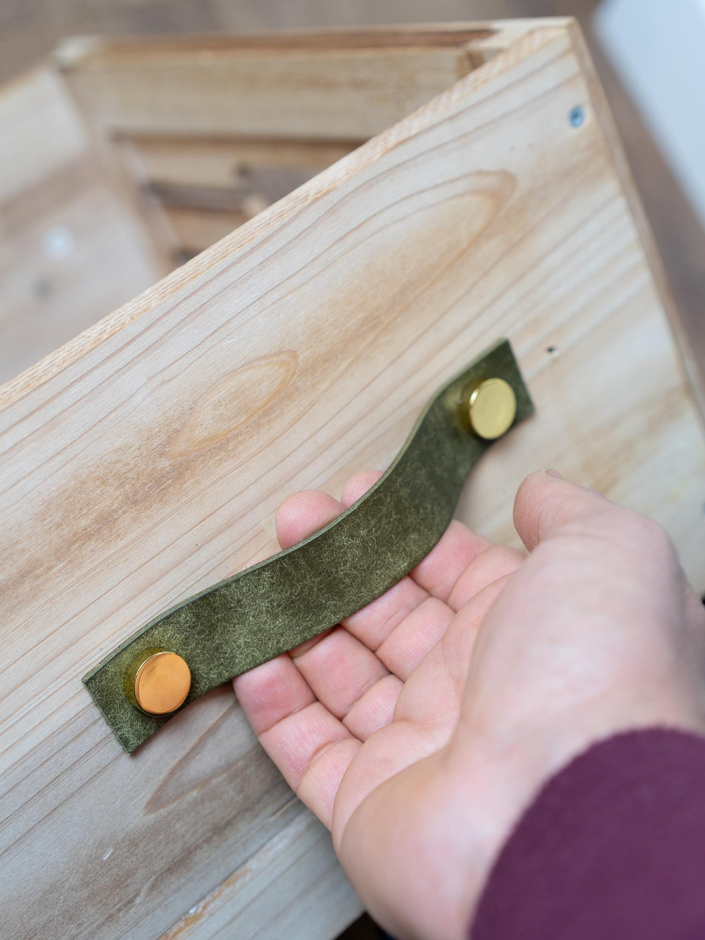 Könnte beinhalten: Nahaufnahme einer Holzkiste mit einem grünen Ledergriff. Der Griff ist mit zwei goldfarbenen Nieten befestigt. Das Holz hat eine helle, natürliche Oberfläche.