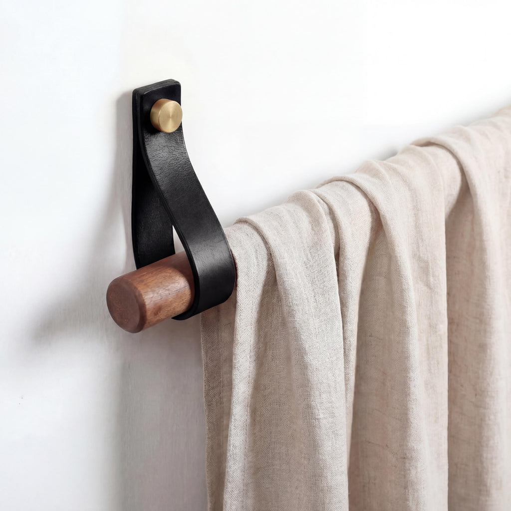 Könnte beinhalten: Ein an der Wand montierter Handtuchhalter aus schwarzem Leder und Holz. Er besteht aus einer dunklen Holzstange, einem schwarzen Lederriemen und einer messingfarbenen Schraube. Ein beiger Stoff hängt an der Halterung vor einer weißen Wand.