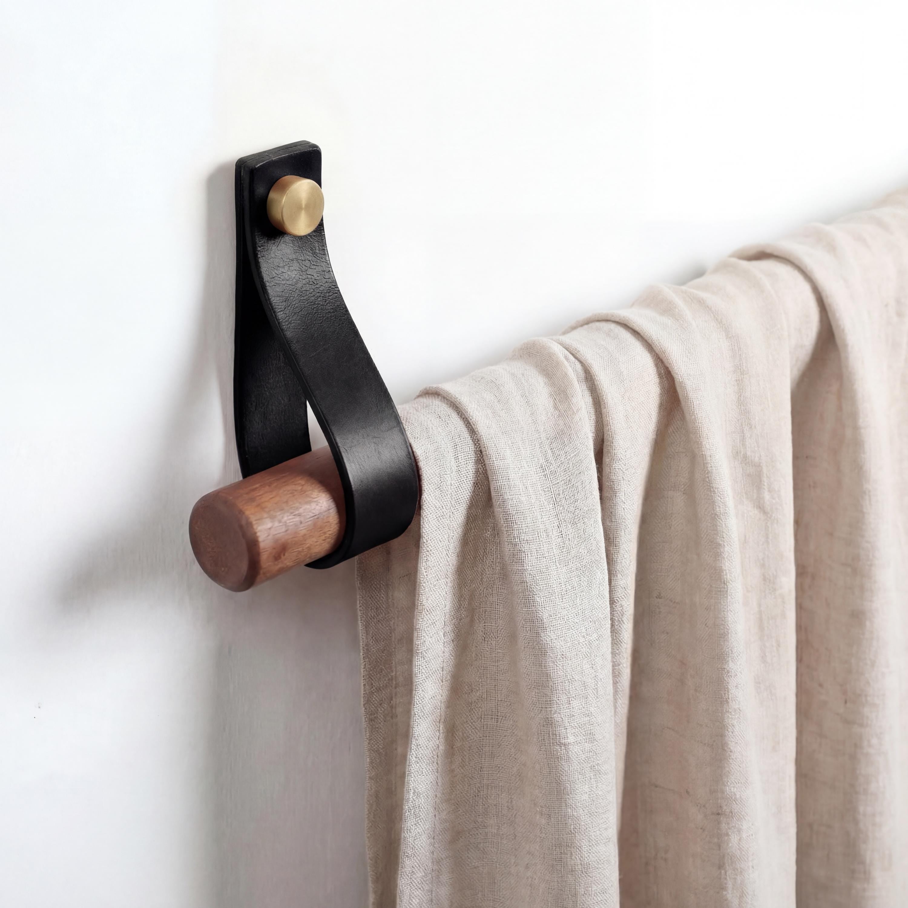 Könnte beinhalten: Ein an der Wand montierter Handtuchhalter aus schwarzem Leder und Holz. Er besteht aus einer dunklen Holzstange, einem schwarzen Lederriemen und einer messingfarbenen Schraube. Ein beiger Stoff hängt an der Halterung vor einer weißen Wand.